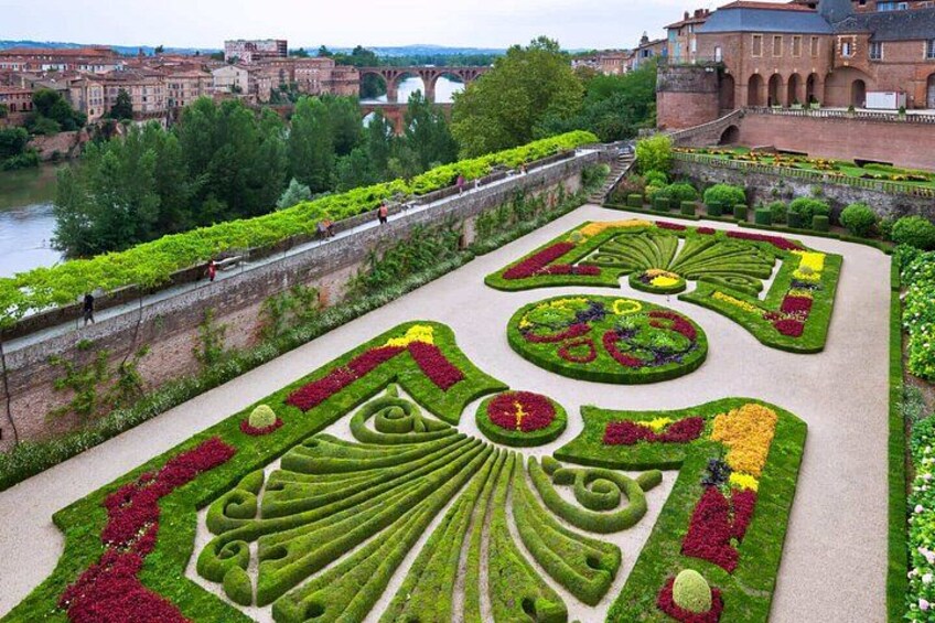 A meticulously landscaped garden with geometric patterns, a river, and historic buildings in the background. 2
