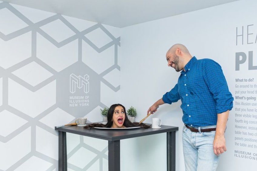 A man in a blue shirt and jeans standing next to a table with a woman sitting at it. The table has a cup and saucer, a small plant, and a book. The background features a geometric wall design and text related to the Museum of Illusions New York. 3