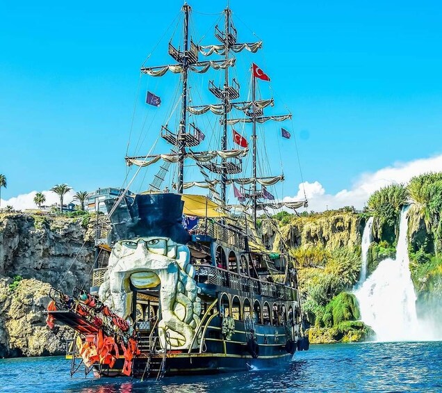 A large ship with multiple masts and flags, docked near a waterfall. 1