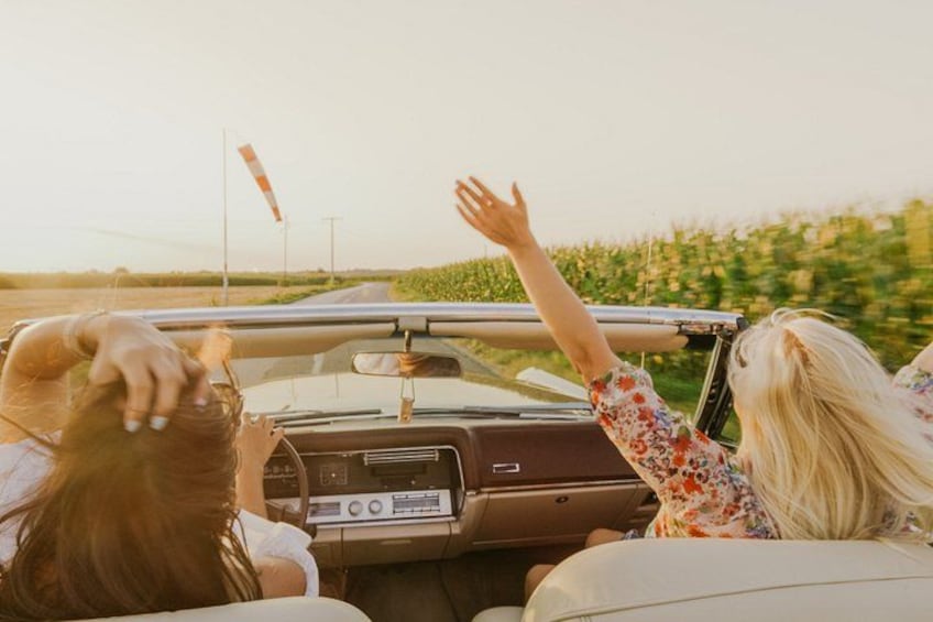Un coche descapotable con dos personas adentro, una de cabello rubio y otra de cabello pelirrojo, conduciendo por una carretera con un campo a un lado. 3