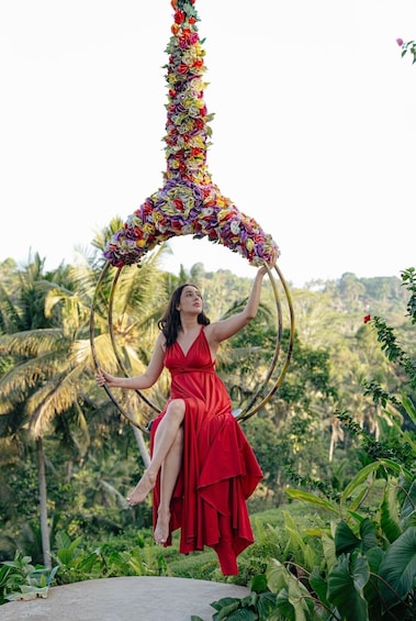 A person in a red dress sitting on a circular structure made of flowers and bamboo, surrounded by lush greenery. 4