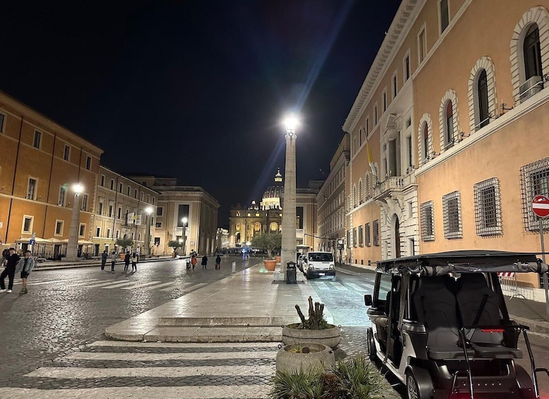 A nighttime street scene with historic buildings, a fountain, and a parked vehicle. 4