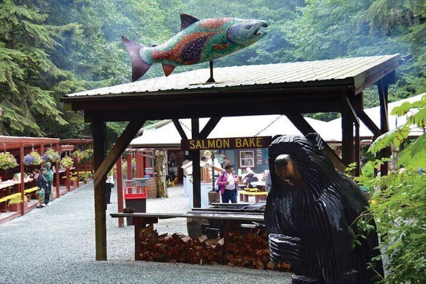 A salmon bake sign, a large fish sculpture, and a bear statue in a forested area. 5