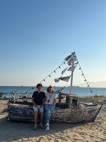 A couple sitting on a beach by a weathered boat with striped sails. 5