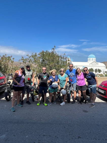 A group of people posing in front of a white building with blue domes, likely a church or historical site, with parked motorcycles and a clear blue sky in the background. 1