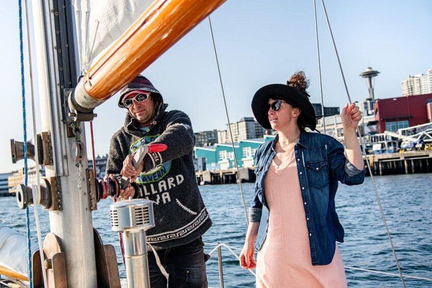 Une personne manœuvrant un treuil sur un bateau, une autre personne se tenant à proximité, et un paysage urbain en arrière-plan. 2