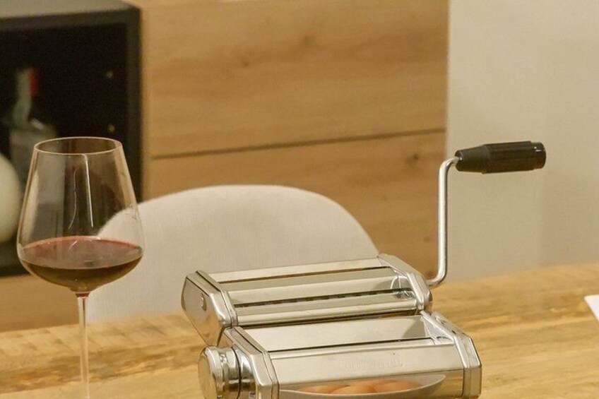 Eine transparente Glas-Pastamachine auf einem Holztisch, daneben ein Glas Rotwein. 3