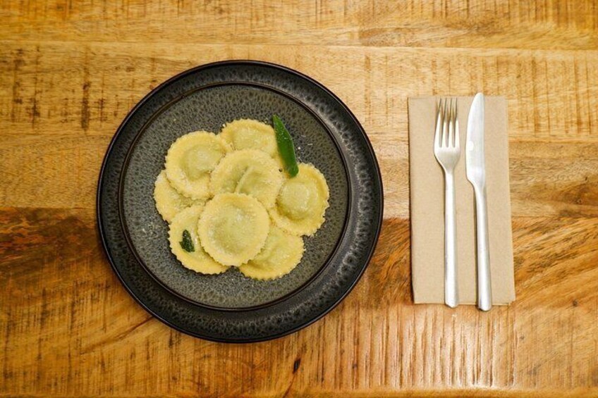 Ein Teller Ravioli mit grüner Dekoration auf einem dunklen Teller, dazu eine Gabel und ein Messer auf einem Holztisch. 1