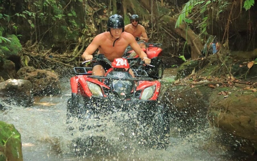 Two people on red ATVs navigating a shallow water crossing in a forested area. 1