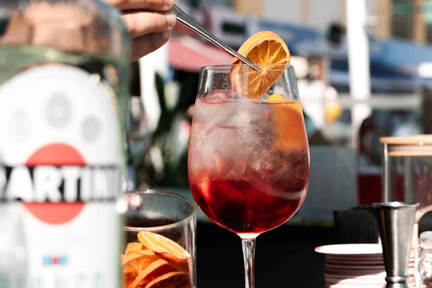 A cocktail glass with ice and an orange slice garnish, a bottle of Martini in the background, and a blurred outdoor setting. 1