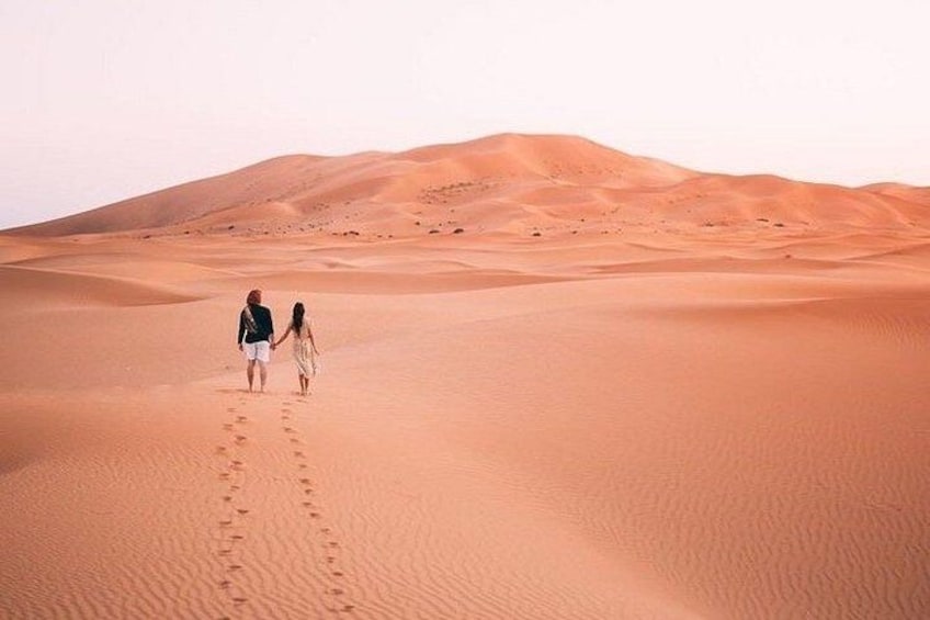 Two people walking on a sandy desert with footprints leading towards a dune. 1