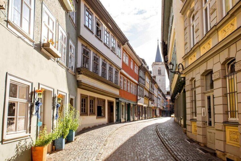 Eine Kopfsteinpflasterstraße gesäumt von historischen Gebäuden, einem Kirchturm und Kübelpflanzen. 4