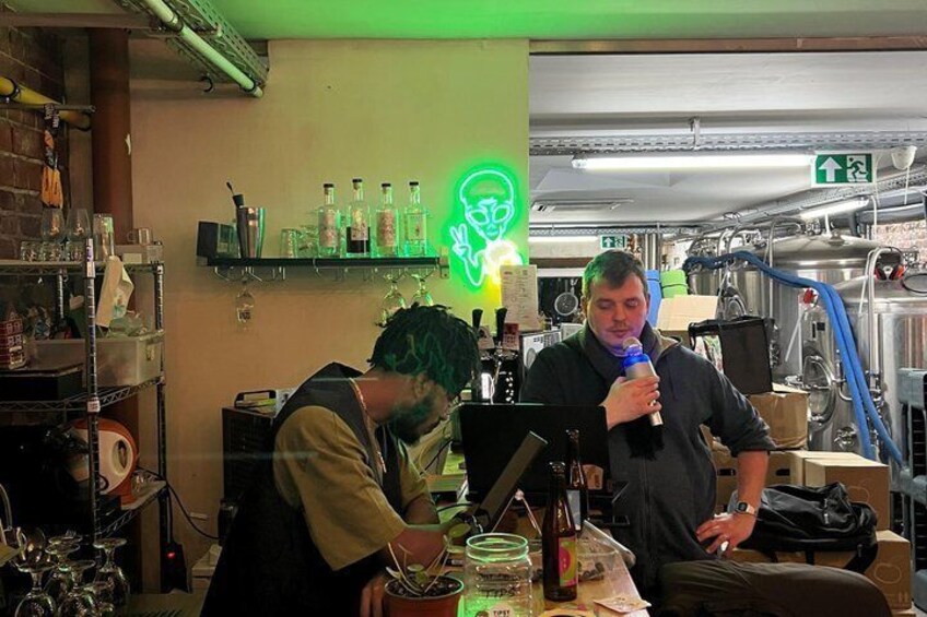 Two people in a bar setting with shelves of bottles and a neon alien sign. 5