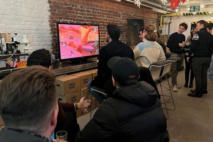 A group of people watching a video on a large screen in a room with brick walls and cardboard boxes labeled 'TIPSY TRIBE' on the floor. 3