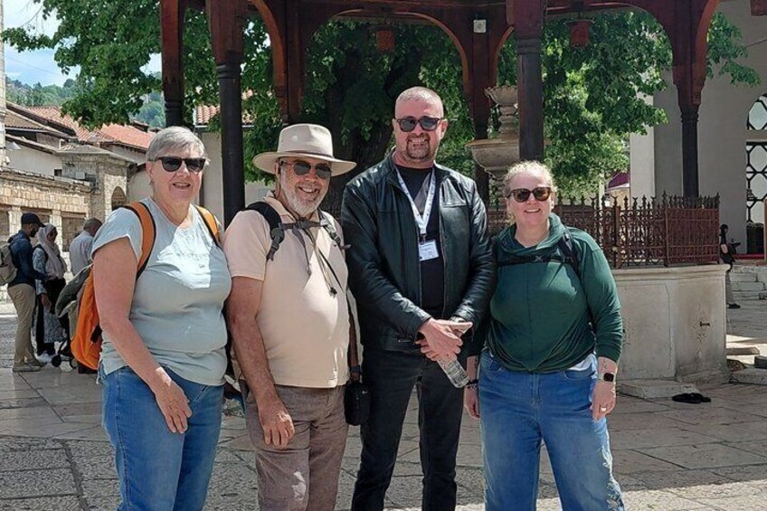Four people standing in a historical square with arches and a fountain. 4