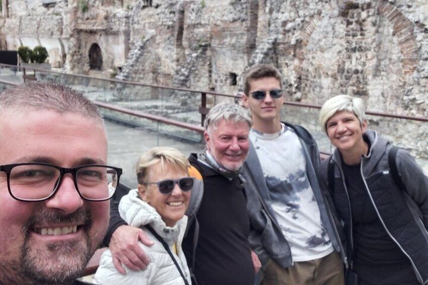 A group of people standing in front of a historical stone structure with arches and a river in the background. 3