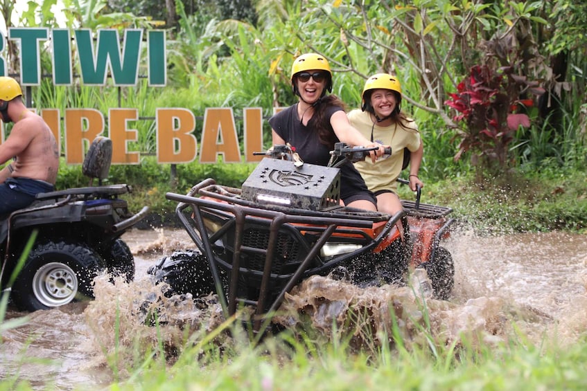 People on an ATV navigating through a flooded area with a sign reading 'TIWI REBAL' in the background. 1