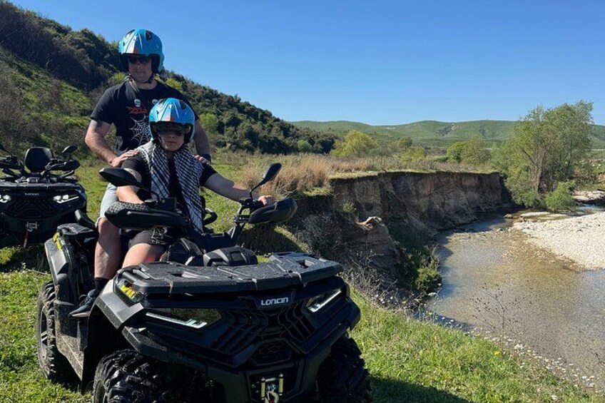 Two people on all-terrain vehicles near a river with a cliff in the background. 4