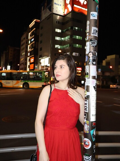 A woman in a red dress standing next to a pole covered with stickers, with a bus and illuminated buildings in the background. 5