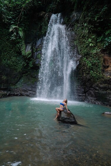 A waterfall in a forested area with clear water and a person sitting on a rock. 5