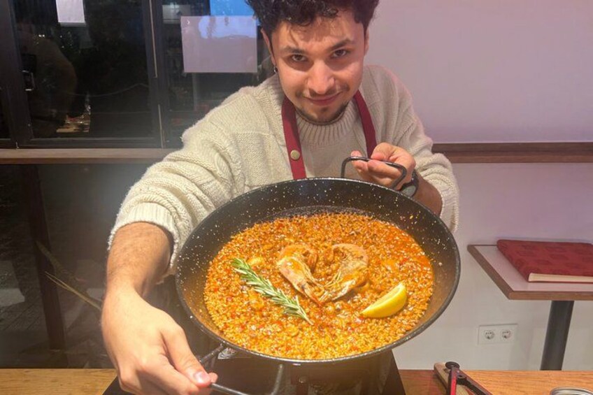 Eine Person hält eine große Pfanne mit einem Gericht, das wie Eintopf oder Curry zubereitet wurde und Fleisch und Gemüse enthält. Darauf liegen ein Zitronenschnitt und ein Rosmarinzweig. 2