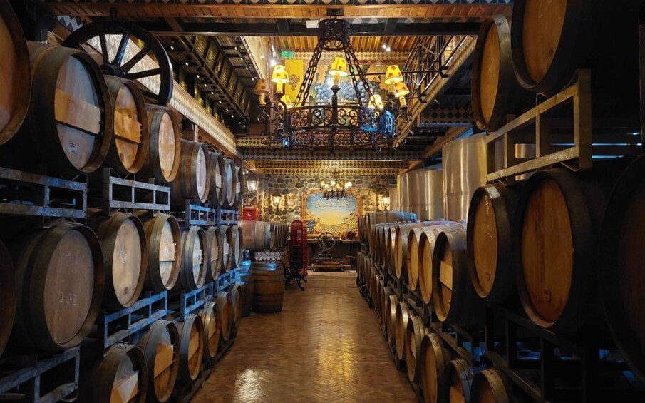 A cellar with wooden barrels stacked on shelves, a chandelier, and a stone wall with a mural. 2
