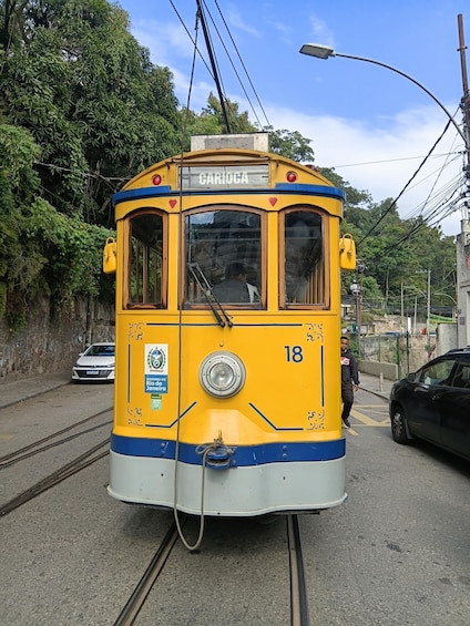 A yellow trolley with 'CARIÓCA' written on it, number 18, on tracks with a streetcar stop sign and a white car in the background. 1