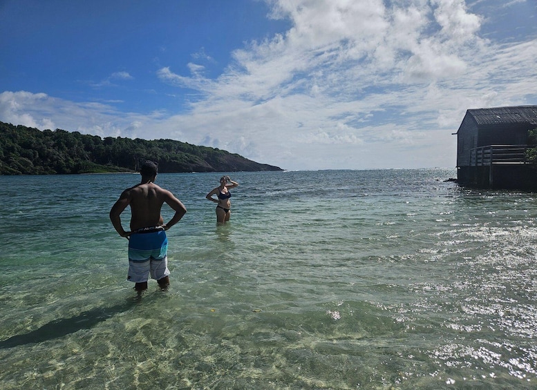 Two people in clear water near a dock, with a forested hill in the background. 1