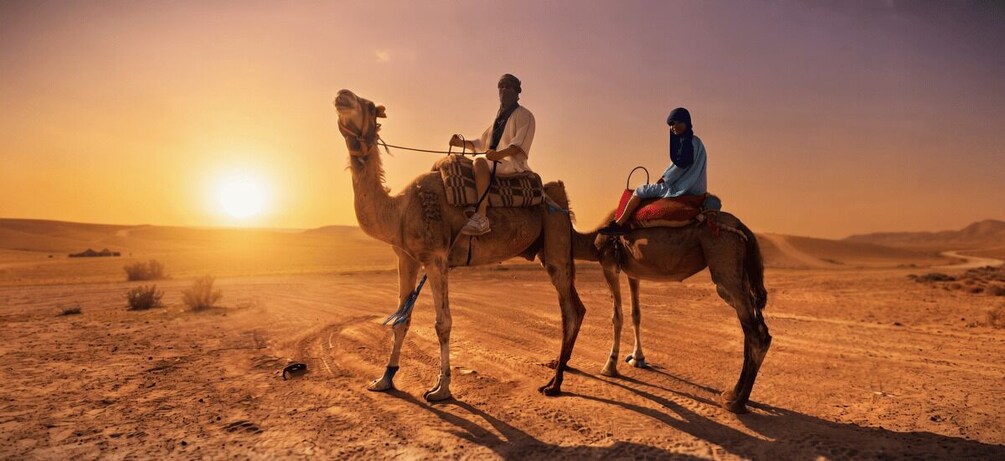 Two people riding camels in a desert landscape during sunset. 2