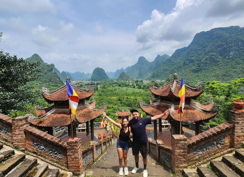 A couple standing in front of traditional architecture with flags, surrounded by lush green mountains. 3