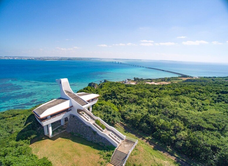 A white building with a unique roof design, surrounded by lush greenery and overlooking a clear blue sea with a long bridge in the distance. 3