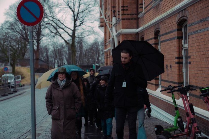 En grupp människor går på en trottoar, några håller i paraplyer, och i närheten står cyklar parkerade. 5
