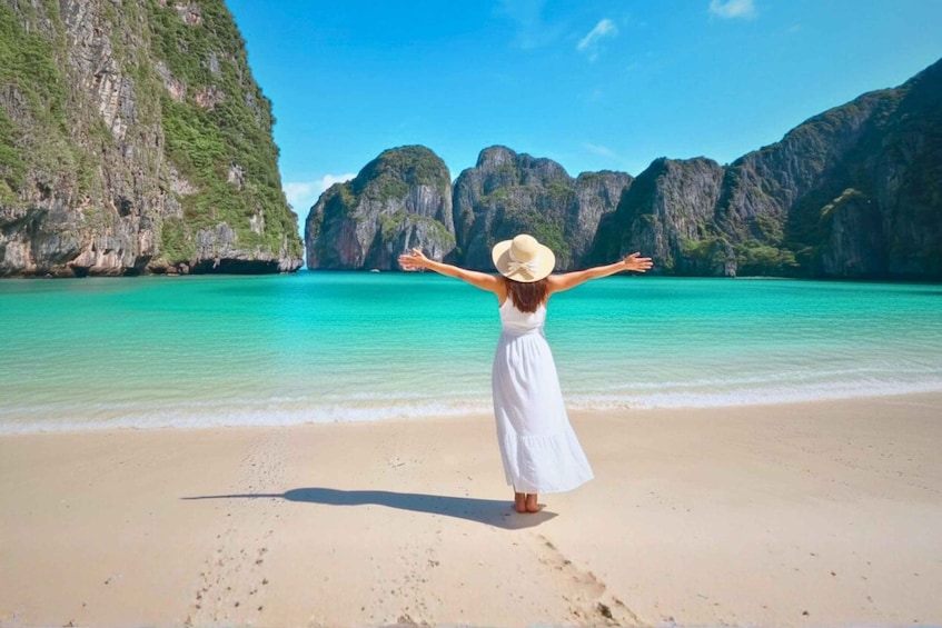 A woman in a white dress and hat standing on a beach with arms outstretched, facing large limestone cliffs. 1