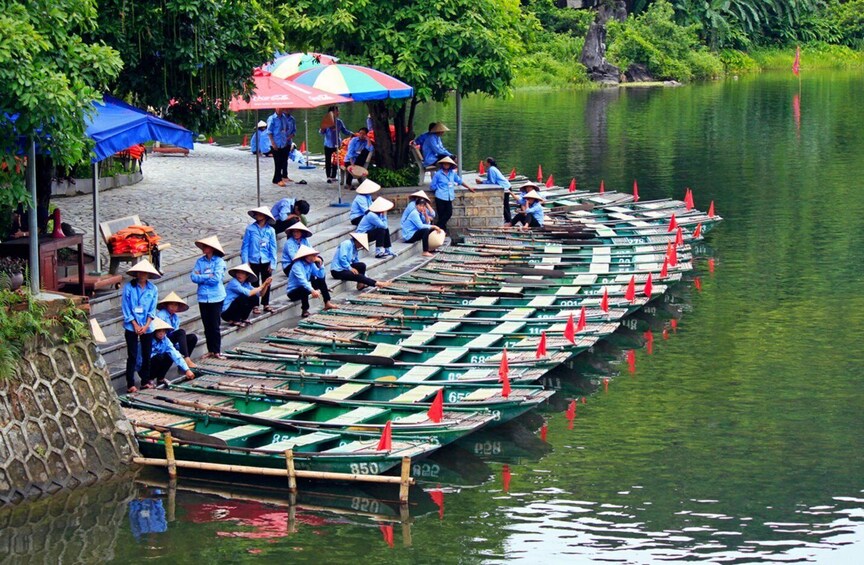 A group of people in blue uniforms and conical hats, seated on a dock by a calm river, with rows of green boats marked with numbers. 2