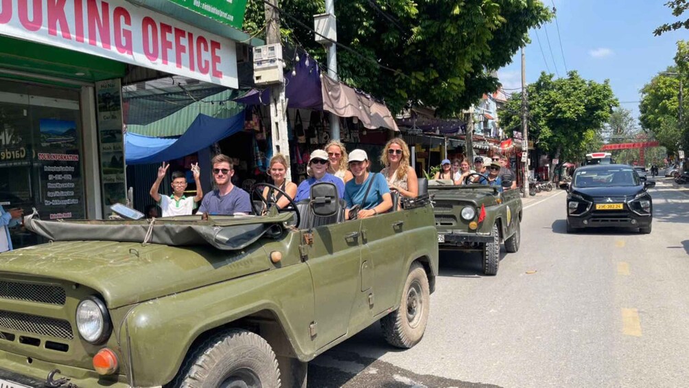 A group of tourists in a green jeep on a street with a sign for a booking office. 4