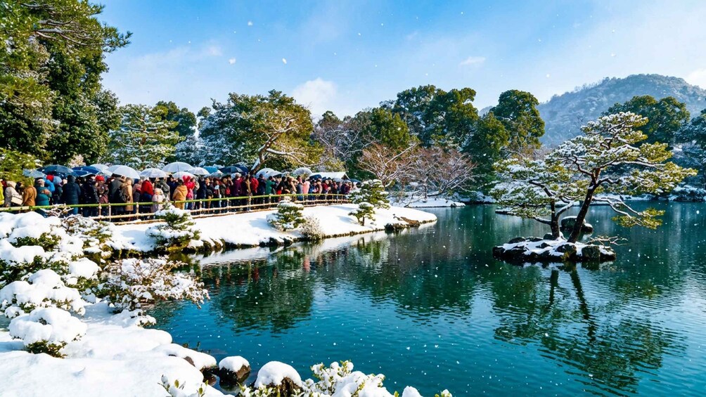 A snowy landscape with a crowd of people standing by a river, snow-covered trees, and a clear blue sky. 4