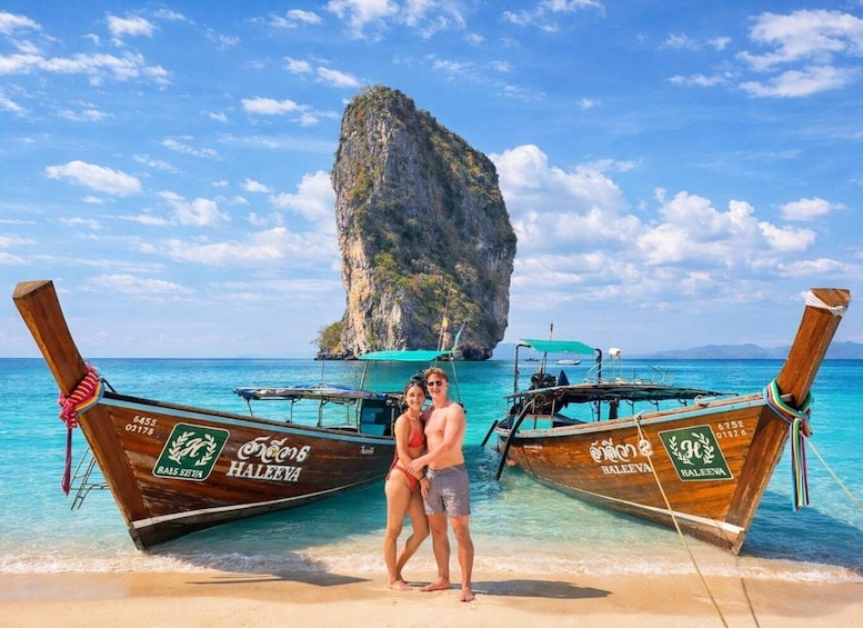 A couple standing between two traditional wooden boats on a beach with clear turquoise water and a prominent limestone rock formation in the background. 3