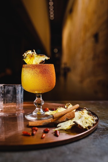 A cocktail in a glass with a frothy top, garnished with a leaf, on a wooden tray with scattered spices and a cinnamon stick. 4