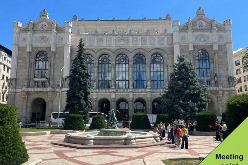 A historic building with ornate architecture, a central fountain, and people gathered around. 1