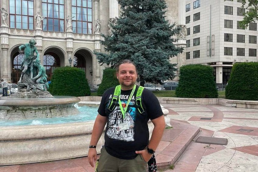 A man standing in front of a fountain with a statue, in an urban square with buildings and a large tree. 5