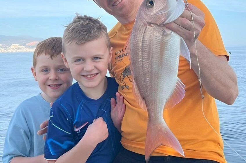 En person holder en fisk, mens to børn står ved siden af, og et vandareal ses i baggrunden. 1