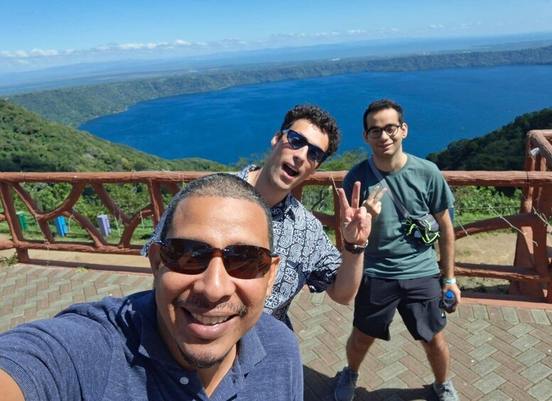 Three people taking a selfie with a scenic view of a lake and hills in the background. 1