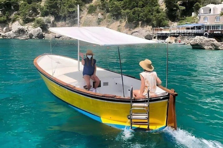 Una barca gialla con due persone a bordo, una in piedi e una seduta, su un mare turchese con una costa rocciosa sullo sfondo. 1