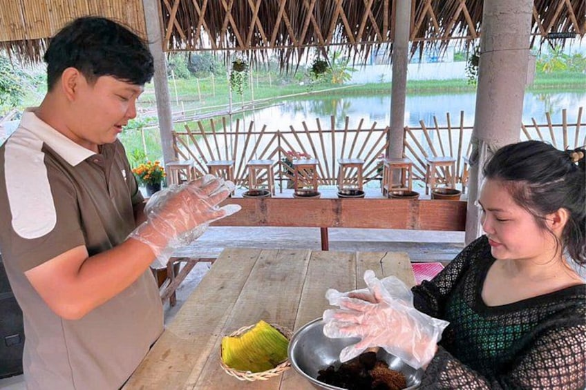 A person preparing food outdoors with a table and chairs, a thatched roof structure, and a body of water in the background. 3