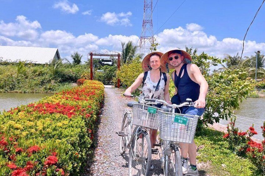 Two people on a bicycle with baskets, a pathway, and a water body with colorful plants. 1