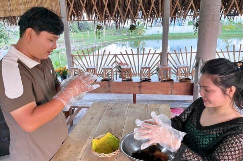 A person in a brown shirt and apron, and another in a black top, both wearing gloves, preparing food at a wooden table outdoors with a thatched roof and a view of a water body. 4