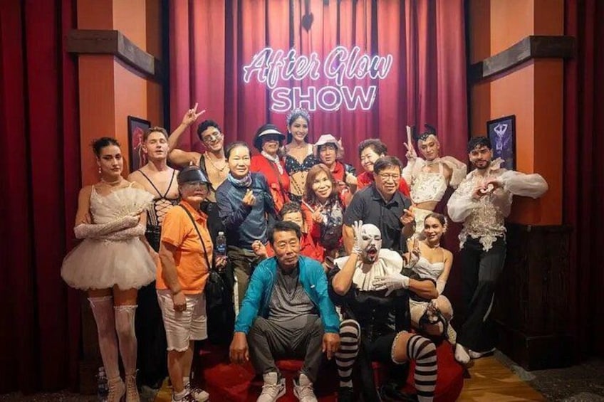 A group of people posing in front of a red curtain with a neon sign that reads 'After Glow SHOW'. 3