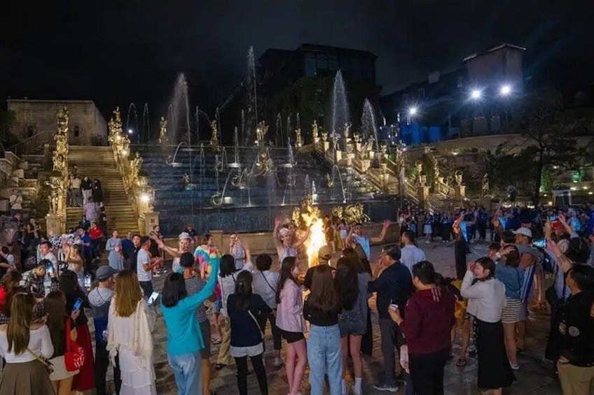 A nighttime gathering of people around a fountain with water jets and illuminated steps. 4