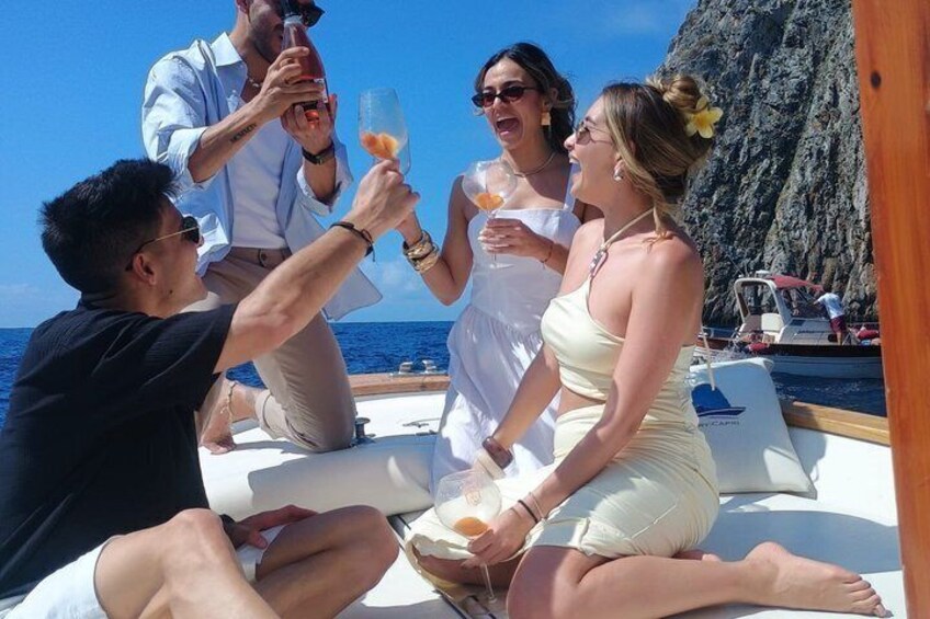 A group of people on a boat, toasting with drinks, with a rocky coastline in the background. 1