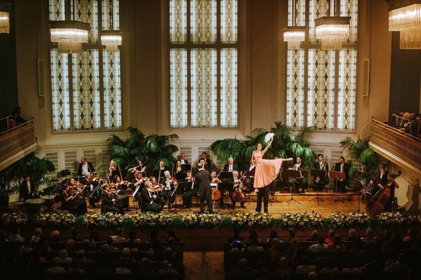Un concierto de música clásica en un salón imponente, con una orquesta extensa, un solista y un público sentado en filas. 1